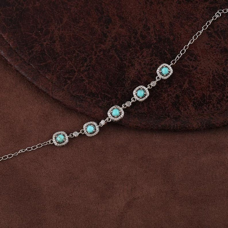 Pode incluir: Uma pulseira de prata com cinco pedras turquesas quadradas, cada uma rodeada por pequenas pedras preciosas transparentes. A pulseira &eacute; exibida em uma superf&iacute;cie marrom, destacando sua delicada corrente e design intrincado.
