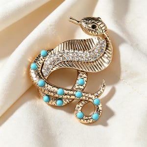 Pode incluir: Um broche de cobra em tom dourado com corpo texturizado, cabeça com um olho preto, uma fileira de strass transparentes brilhantes no capuz e vários cabochões redondos de cor turquesa incrustados ao longo do corpo enrolado.