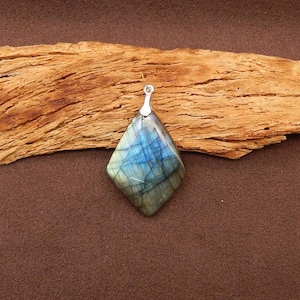 May include: A diamond-shaped pendant with a silver bail. The pendant is made of a blue and green iridescent stone, with a pattern of blue lines. The pendant is displayed on a piece of wood.