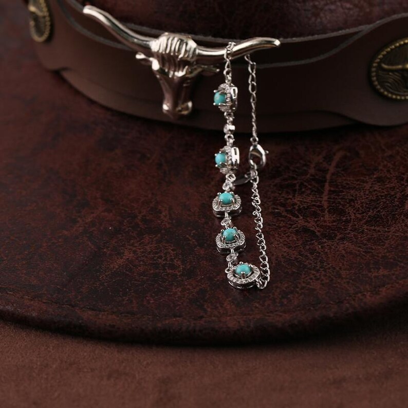 Pode incluir: Um acess&oacute;rio de chap&eacute;u prateado com pedras de cor turquesa e uma corrente, pendurado em um chap&eacute;u de cowboy de couro marrom. O acess&oacute;rio apresenta um design de cabe&ccedil;a de touro.