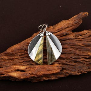 Könnte beinhalten: Silberne Ohrringe mit einem einzigartigen Design. Jeder Ohrring hat eine Tropfenform mit einer Kombination aus dunkelgrauem Metall, Weiß und schillerndem grün-goldenem Stein. Die Ohrringe werden auf einem Holzstück präsentiert.