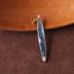May include: A clear, elongated pendant with blue and silver inclusions, suspended from a small gold-colored loop. The pendant has a pointed tip and a rough, textured appearance. The background is a mix of brown and dark brown.