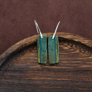 Op de afbeelding: Een paar rechthoekige oorbellen met zilveren haken. De oorbellen zijn gemaakt van een steen met blauwe, groene en gele tinten. Ze worden gepresenteerd op een houten oppervlak.