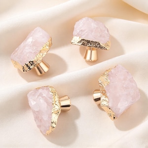 May include: Four cabinet knobs featuring irregular, raw-cut pale pink rose quartz stones. Each stone is accented with a metallic gold-toned finish along the edges and is mounted on a polished gold-toned cylindrical metal base.