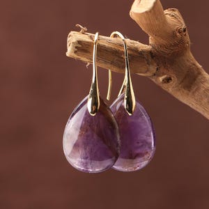 May include: A pair of teardrop earrings with gold-coloured hooks. The earrings have a translucent, deep purple stone with lighter purple and brown inclusions. They are suspended from a wooden branch against a brown backdrop.