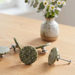 May include: A set of round cabinet knobs featuring a mottled olive green stone surface with a polished finish. Each knob is attached to a silver-toned metal base and includes a threaded screw for installation, showcasing a natural speckled texture.