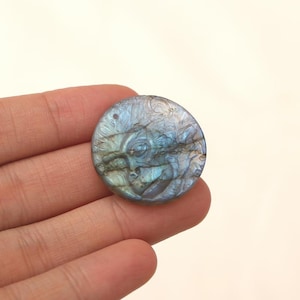 May include: A round, carved gemstone pendant featuring a face with detailed features. The stone has a mix of blue, gray, and iridescent colors. A small hole is drilled at the top for stringing. The pendant is held in a hand.