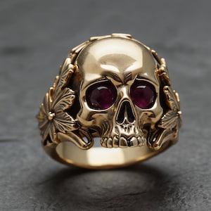 May include: A gold-toned ring featuring a central skull design with deep red, faceted gemstone eyes. The sides of the ring are decorated with detailed, raised leaf patterns that extend from the skull, creating a cohesive, ornate metal band.