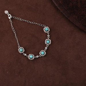 Pode incluir: Uma pulseira de prata com pedras quadradas turquesa, cada uma rodeada por pequenos cristais transparentes. A pulseira est&aacute; sobre uma superf&iacute;cie castanha.