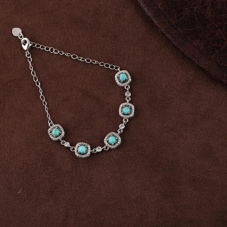 Pode incluir: Uma pulseira de prata com pedras quadradas turquesa, cada uma rodeada por pequenos cristais transparentes. A pulseira est&aacute; sobre uma superf&iacute;cie castanha.