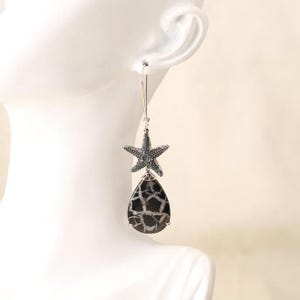 May include: A silver-toned dangle earring featuring a textured, five-pointed starfish charm suspended from a thin wire hook. Below the starfish hangs a smooth, teardrop-shaped stone with a dark gray base and a distinct white webbed pattern.