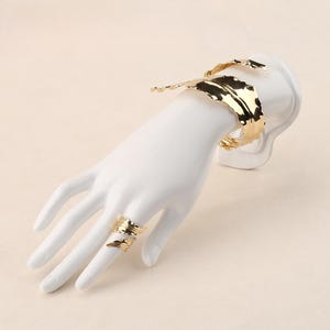 May include: A gold-coloured cuff bracelet and ring set. The cuff bracelet features a leaf-like design, and the ring has a similar, smaller leaf-like pattern. The jewellery is displayed on a white hand-shaped mannequin, set against a neutral background.