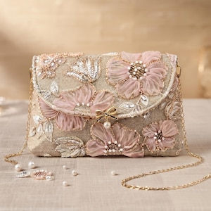 May include: A rectangular clutch bag in a champagne-coloured fabric, featuring three-dimensional pink organza flowers, silver sequin embroidery, and pearl accents. The bag includes a gold-tone metal chain strap and a small gold bow clasp with a pearl.