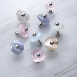 May include: A collection of eight round cabinet knobs featuring smooth, translucent, cat-eye style glass in various pastel colours including white, pink, blue, and beige, each centred with a small, polished silver-tone metal stud and base.