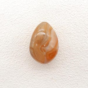 May include: A polished, teardrop-shaped gemstone with a swirling pattern of brown, orange, and cream colors. The stone has a glossy finish and is set against a white background. The gemstone is likely used for jewelry or decorative purposes.