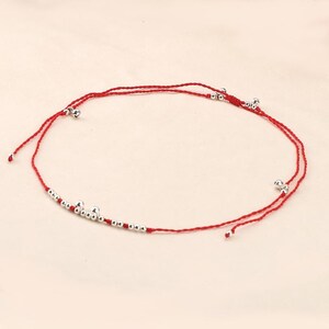 May include: Red string anklet with silver-colored beads and small bells. The anklet features a simple design with the beads and bells evenly spaced along the red string. A delicate piece of jewelry.