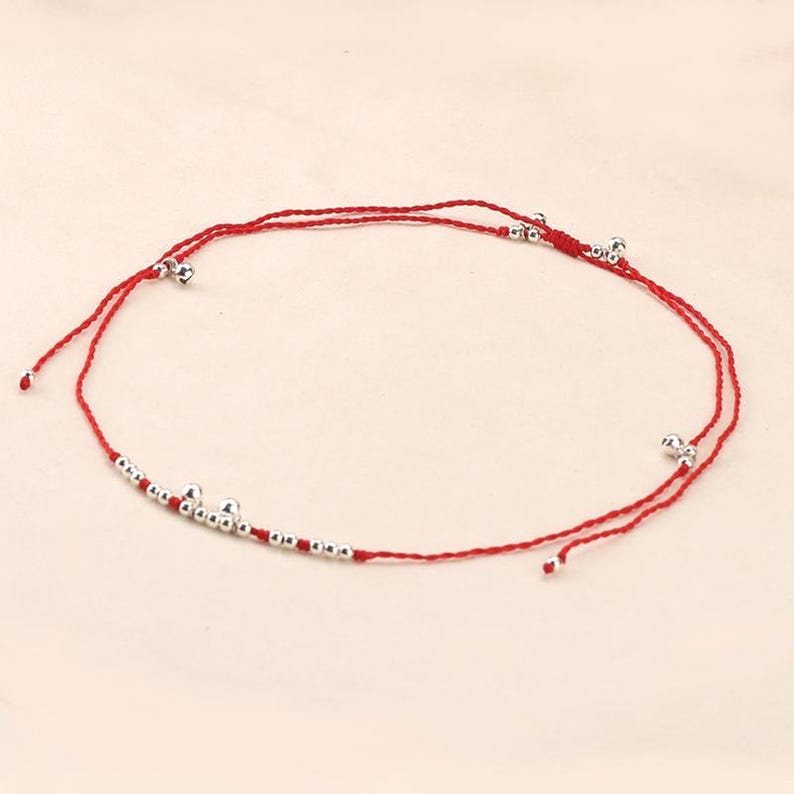 May include: Red string anklet with silver-colored beads and small bells. The anklet features a simple design with the beads and bells evenly spaced along the red string. A delicate piece of jewelry.