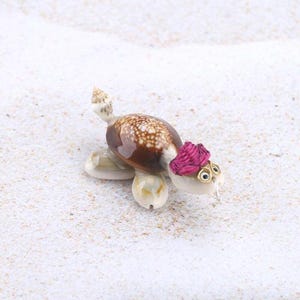 May include: A small turtle figurine crafted from natural seashells. The shell features a brown and white spotted pattern, while the head is adorned with a bright pink coiled fibre accent and small gold-toned wire glasses with black eyes.