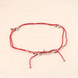 May include: Red string anklet with silver-colored beads. The anklet features a simple design with multiple small, round beads evenly spaced along the red string. The anklet is tied with a knot at the end.
