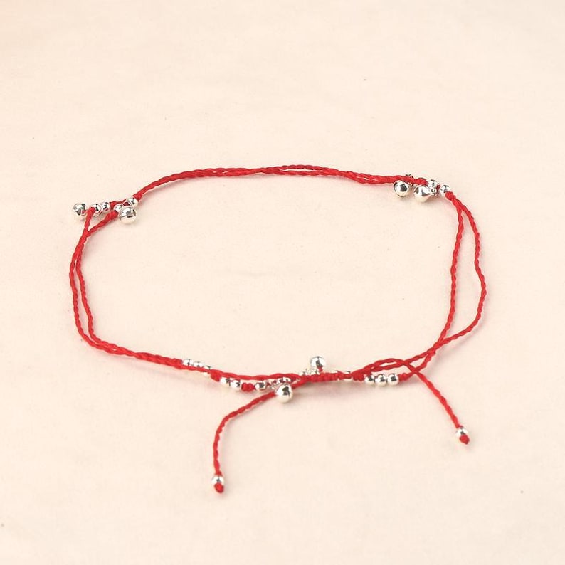 May include: Red string anklet with silver-colored beads. The anklet features a simple design with multiple small, round beads evenly spaced along the red string. The anklet is tied with a knot at the end.