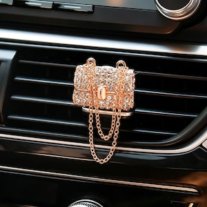 Può includere: Un deodorante per auto a forma di borsa in miniatura, ricoperto di strass trasparenti scintillanti con una cornice in metallo dorato e una tracolla a catena dorata pendente, fissato su una bocchetta di ventilazione nera orizzontale.