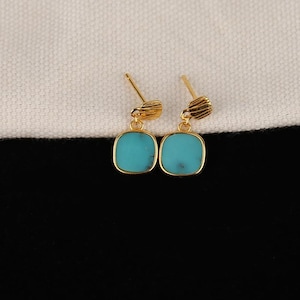 Può includere: Un paio di orecchini a bottone color oro con pietre quadrate color turchese. Gli orecchini hanno un design strutturato nella parte superiore. Sono esposti su uno sfondo bianco e nero.