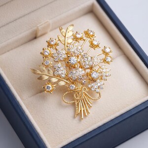 Op de afbeelding: Een goudkleurige broche in de vorm van een boeket bloemen, voorzien van meerdere heldere, fonkelende stenen in de bloemblaadjes en harten van de bloemen. Het ontwerp bevat slanke gouden bladeren en een gestrikt lintdetail aan de onderkant.