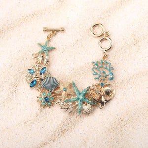 May include: Gold-coloured bracelet with marine-themed charms. It features turquoise starfish, coral, and seashell details, plus blue crystal accents. The toggle clasp and connecting rings are also gold.
