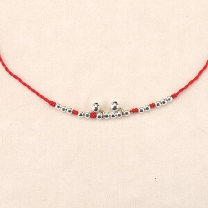 May include: A delicate red string bracelet featuring small silver beads and two silver bells. The bracelet is designed with a simple, elegant style, perfect for everyday wear. The beads and bells add a touch of sparkle to the wrist.
