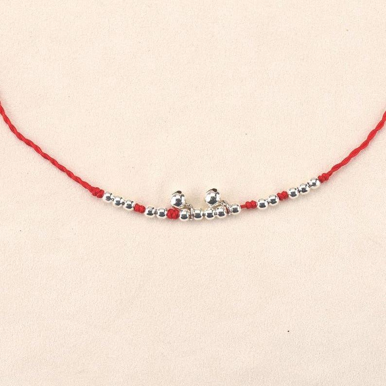 May include: A delicate red string bracelet featuring small silver beads and two silver bells. The bracelet is designed with a simple, elegant style, perfect for everyday wear. The beads and bells add a touch of sparkle to the wrist.