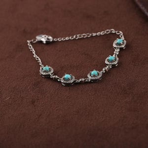 Pode incluir: Uma pulseira de prata com cinco pedras preciosas turquesa, cada uma rodeada por pequenas pedras transparentes. A pulseira tem uma corrente delicada e um fecho. A joia &eacute; exibida numa superf&iacute;cie castanha.