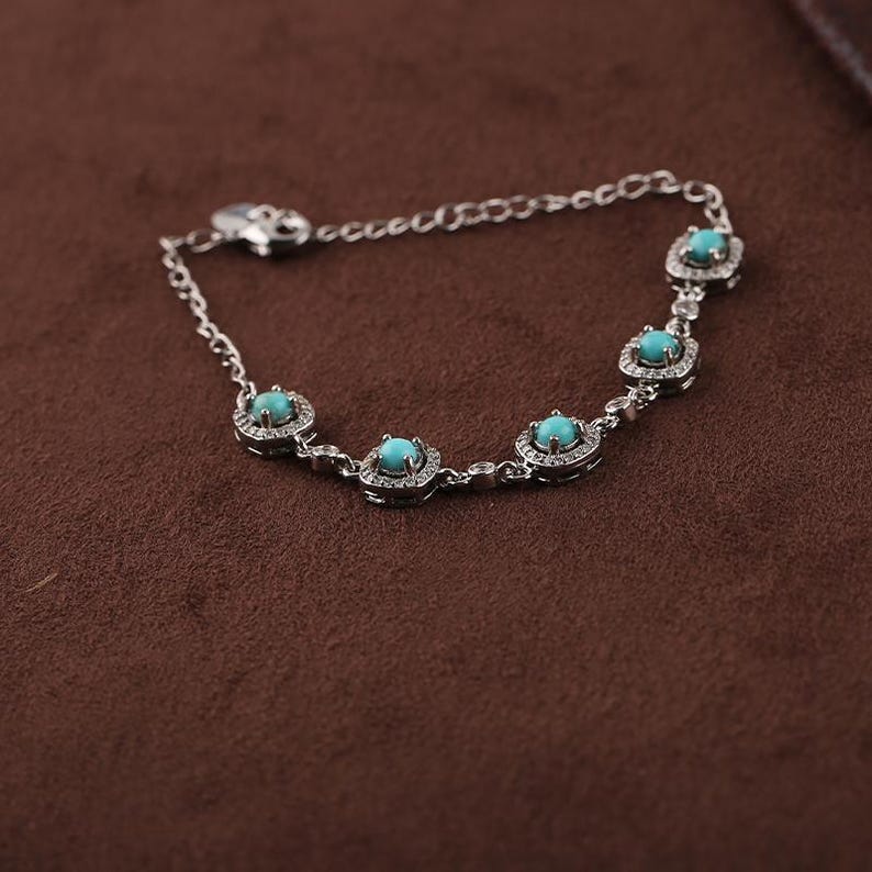 Pode incluir: Uma pulseira de prata com cinco pedras preciosas turquesa, cada uma rodeada por pequenas pedras transparentes. A pulseira tem uma corrente delicada e um fecho. A joia &eacute; exibida numa superf&iacute;cie castanha.
