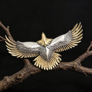 Pode incluir: Um ornamento de metal em forma de águia com corpo e cabeça em tom prateado polido, contrastando com pontas das asas e penas da cauda em tom dourado. A águia é representada com as asas totalmente abertas em uma posição de voo.