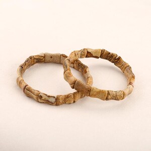 Puede incluir: Dos pulseras de piedra natural. Cada pulsera está hecha de cuentas rectangulares de color marrón claro con vetas marrones más oscuras. Las pulseras son circulares y descansan sobre una superficie blanca.