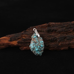 May include: A turquoise stone pendant with a silver bail. The pendant has a rough, natural texture with a mix of brown and blue-green colors. The stone is irregularly shaped and appears to be a raw, uncut gemstone.