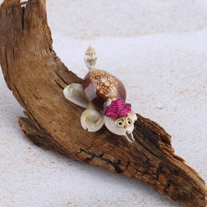 Peut inclure: Figurine de tortue faite à la main en coquillage, avec une carapace marron et blanche, ornée d'une fleur rose et de petites lunettes dorées. La tortue est posée sur du bois flotté.
