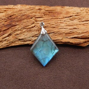 May include: A diamond-shaped labradorite pendant with a silver-colored bail. The gemstone displays iridescent blue and green hues. The pendant is displayed on a piece of textured wood.