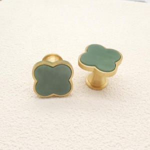 Könnte beinhalten: Zwei dekorative Schrankknöpfe mit Kleeblatt-Design. Die Knöpfe haben ein gedämpftes grünes Kleeblatt, das in einem goldfarbenen Metallrahmen eingefasst ist. Einer ist flach, der andere auf einem kurzen, zylindrischen Sockel montiert.