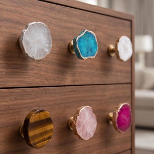 Puede incluir: Seis pomos circulares para armarios fijados en una cómoda de madera. Cada pomo presenta una rodaja de piedra natural única con un borde metálico, incluyendo variedades en blanco, azul, ojo de tigre marrón, rosa pálido y magenta vibrante.