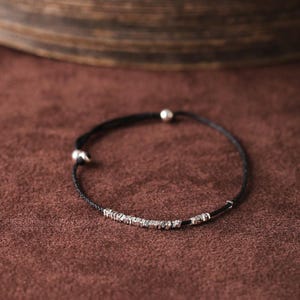 May include: A delicate black cord bracelet featuring a row of small, irregularly shaped silver beads. The bracelet has a simple design with a small silver bead at each end. The background is a solid, warm brown color.