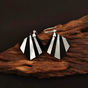 May include: A pair of geometric earrings with a black and white striped design. The earrings are square-shaped with a silver hook. The earrings are displayed on a piece of dark brown wood.