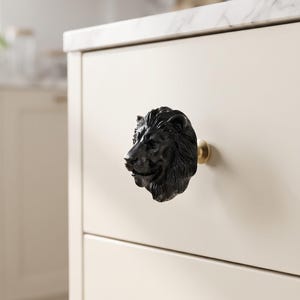 Peut inclure: Un bouton de placard noir mat en forme de tête de lion détaillée, présentant une crinière et des traits du visage sculptés. La tête de lion est fixée sur une base métallique circulaire de couleur laiton, montée sur un tiroir de placard clair.