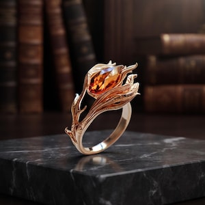 May include: A rose gold ring featuring a marquise-cut amber gemstone set within a floral design. The band is crafted to resemble a stem with textured leaves and thorns, wrapping around the central orange stone in a detailed botanical arrangement.