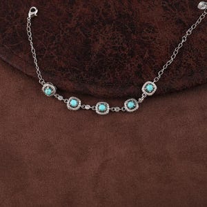 Pode incluir: Uma pulseira de prata com pedras de cor turquesa emolduradas em quadrados, rodeadas por pequenas pedras transparentes. A pulseira tem uma corrente delicada e um fecho de fecho. A joia &eacute; exibida numa superf&iacute;cie castanha.