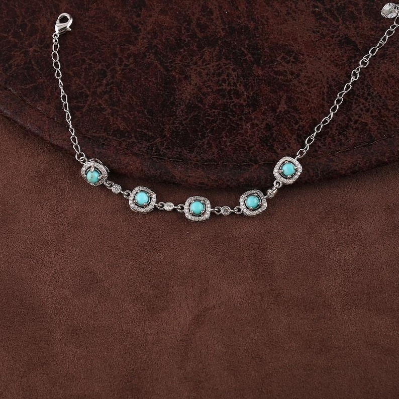 Pode incluir: Uma pulseira de prata com pedras de cor turquesa emolduradas em quadrados, rodeadas por pequenas pedras transparentes. A pulseira tem uma corrente delicada e um fecho de fecho. A joia &eacute; exibida numa superf&iacute;cie castanha.