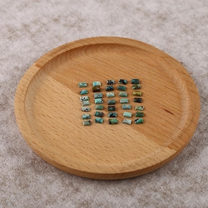 Puede incluir: Un conjunto de gemas rectangulares de turquesa con patrones oscuros se exhiben en una bandeja redonda de madera. Las gemas varían en tonos de azul y verde. La bandeja tiene vetas de madera natural.
