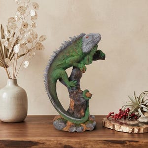 Puede incluir: Una figura de iguana verde posada sobre una base de rama de árbol marrón texturizada. El lagarto tiene la cabeza gris, ojos naranjas y una cola larga que se curva alrededor de la base. La escultura está detallada con escamas y espinas.