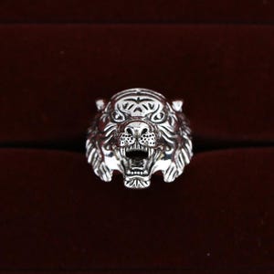 Könnte beinhalten: Ein silberner Ring mit einem detaillierten Tigerkopf-Design. Der Ring zeigt das Gesicht des Tigers mit offenem Maul, Zähnen und komplizierten Fell-Details. Der Ring ist vor einem dunkelroten Hintergrund platziert.
