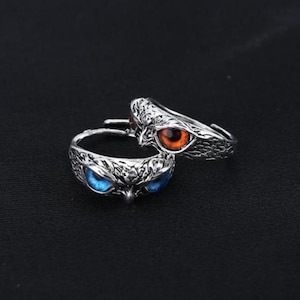 May include: Two silver-toned owl rings with detailed feather textures. One ring features bright orange eyes, the other has striking blue eyes. The rings are set against a dark background, highlighting the intricate design.