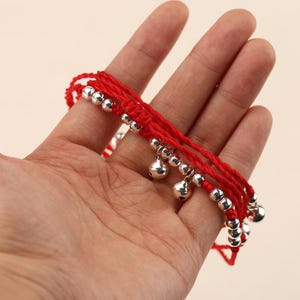 May include: Red anklet with silver-colored beads and small bells. The anklet is made of woven red thread and features multiple strands. The bells and beads are evenly spaced along the anklet.